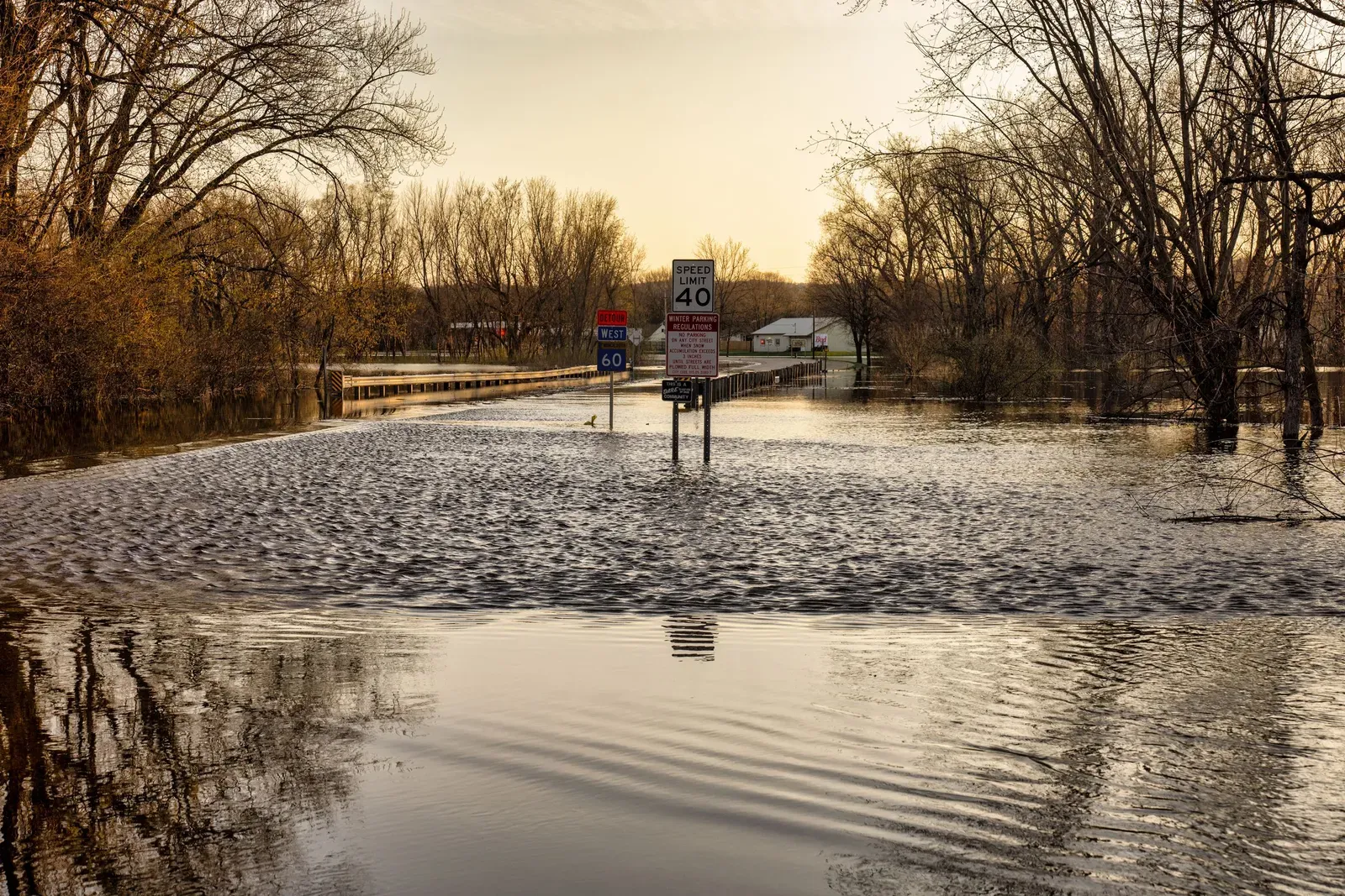 A flooded rural road at dusk with a speed limit sign and route markers partly submerged in floodwater