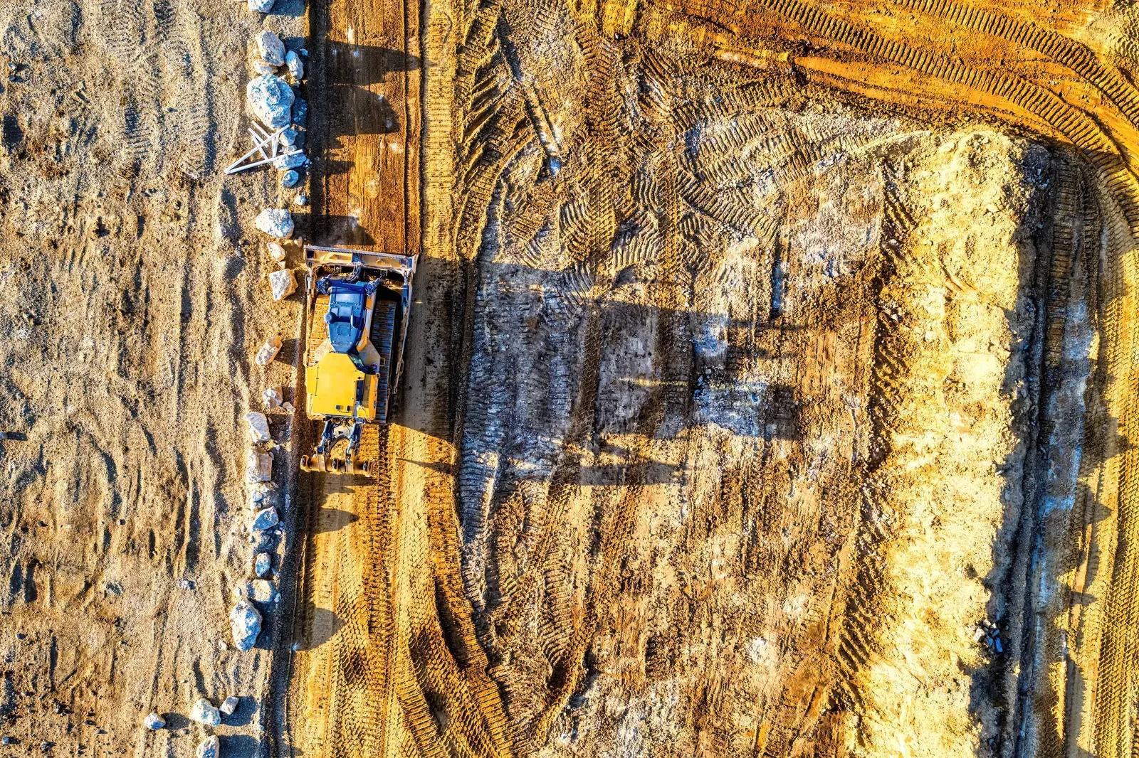 A direct overhead view of a bulldozer shaping a dirt roadbed, with tire tracks curving across bare earth