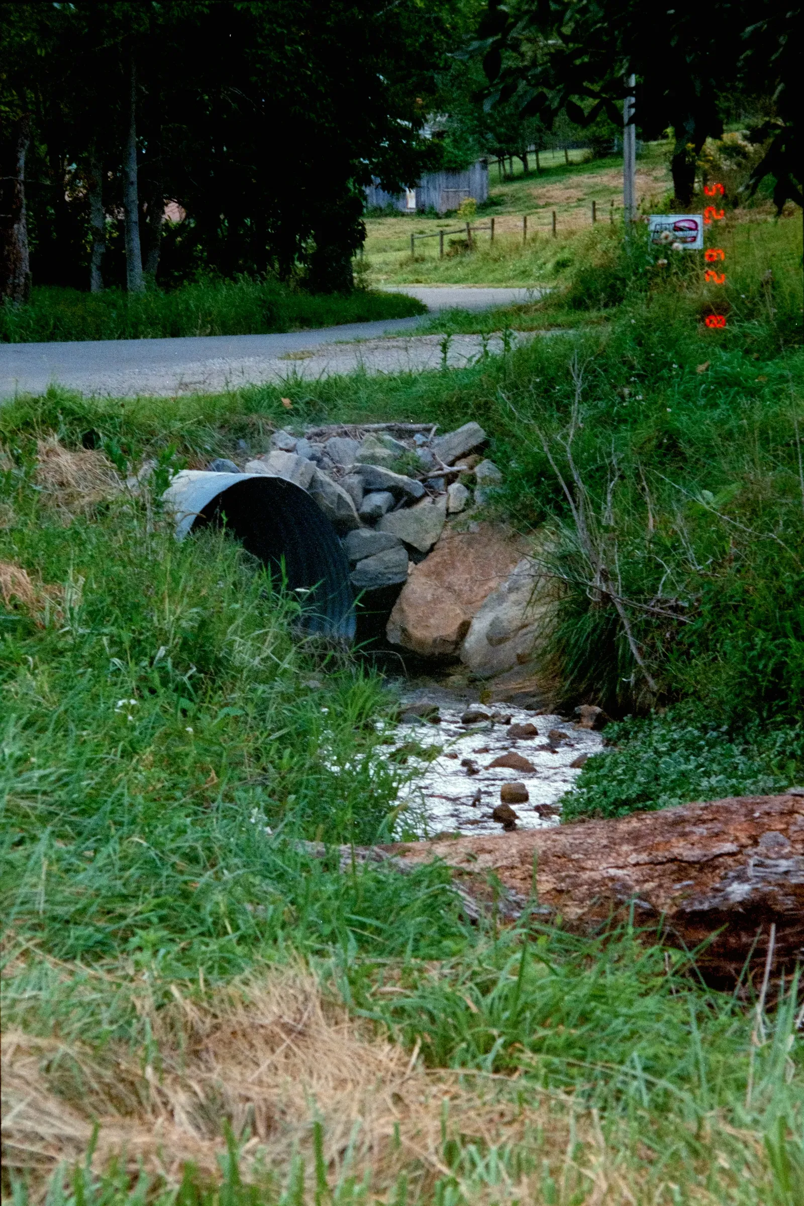 A corrugated metal culvert channeling a small creek beneath a rural gravel road, framed by grass and rock