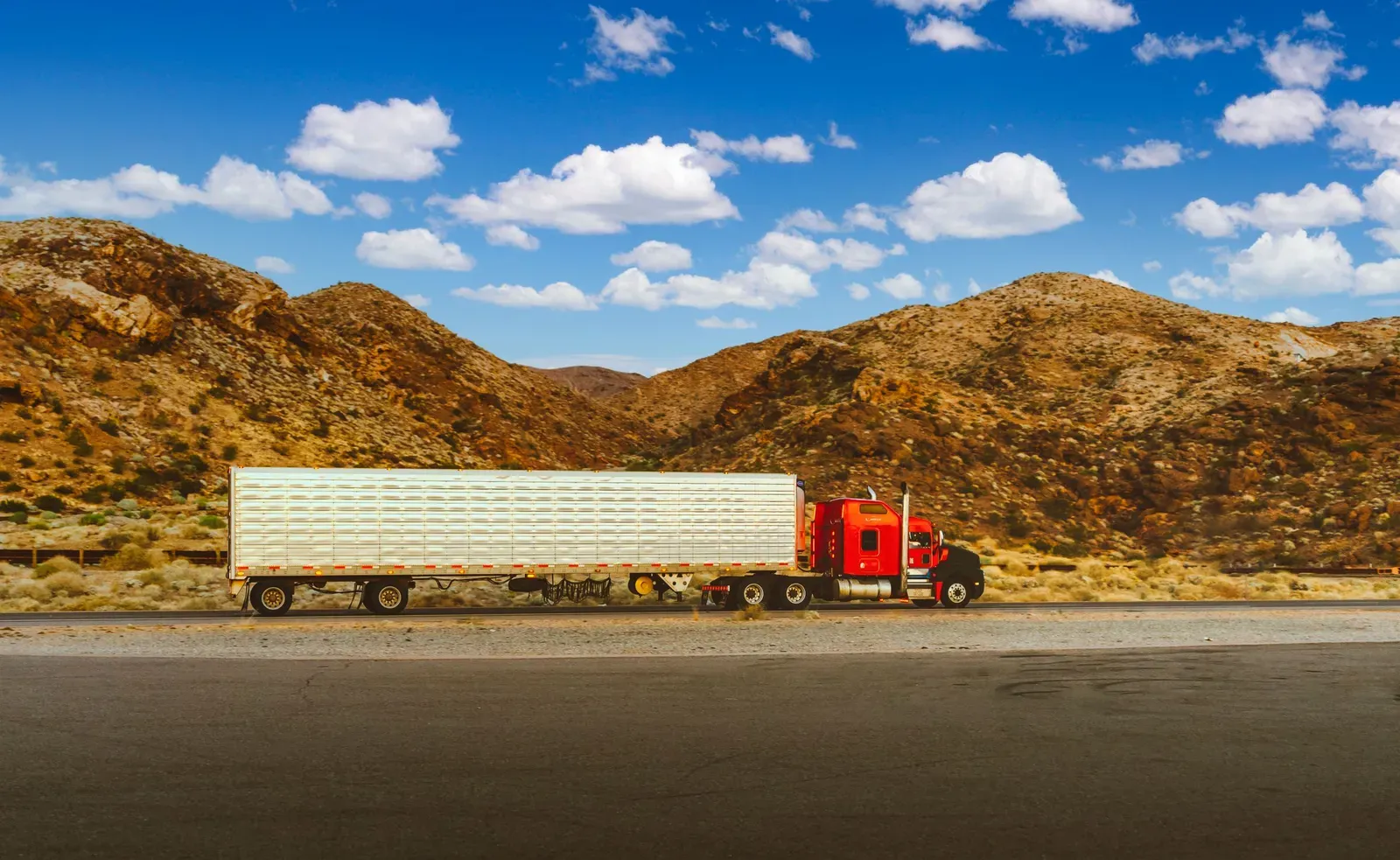 A red semi-truck with a reefer trailer traveling a desert highway with rocky hills behind it