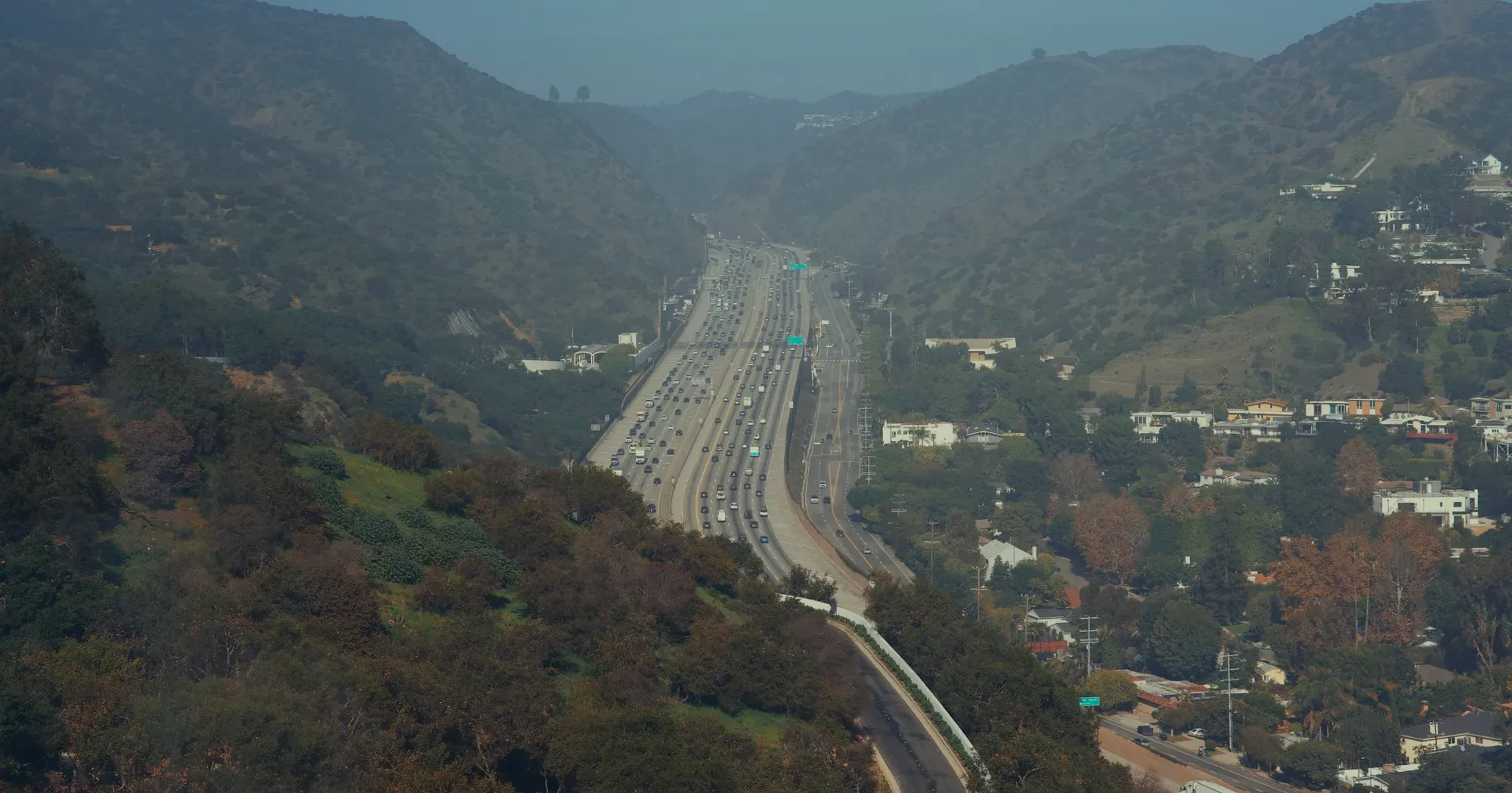 A multi-lane interstate curving through a hazy, tree-covered mountain pass with houses on the hillside beyond