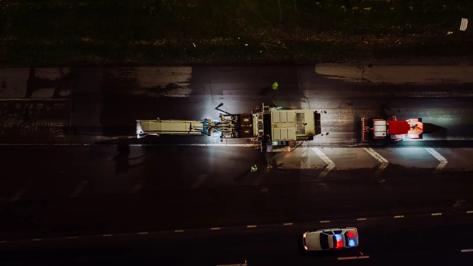 A nighttime aerial view of an asphalt paver and roller laying fresh pavement on a highway lane, lit by work lights