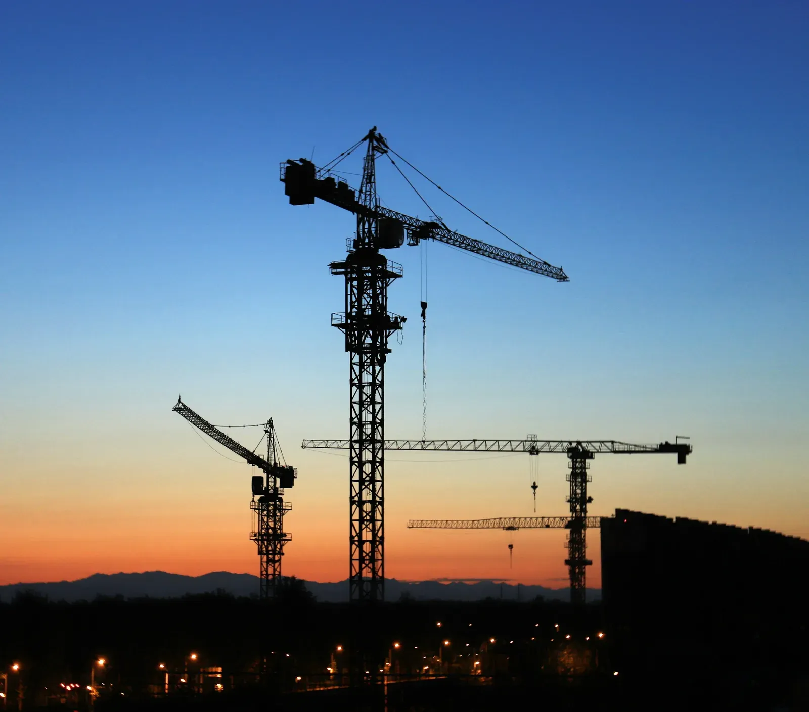 Silhouettes of several tower cranes on a construction site against an orange and blue sunset sky