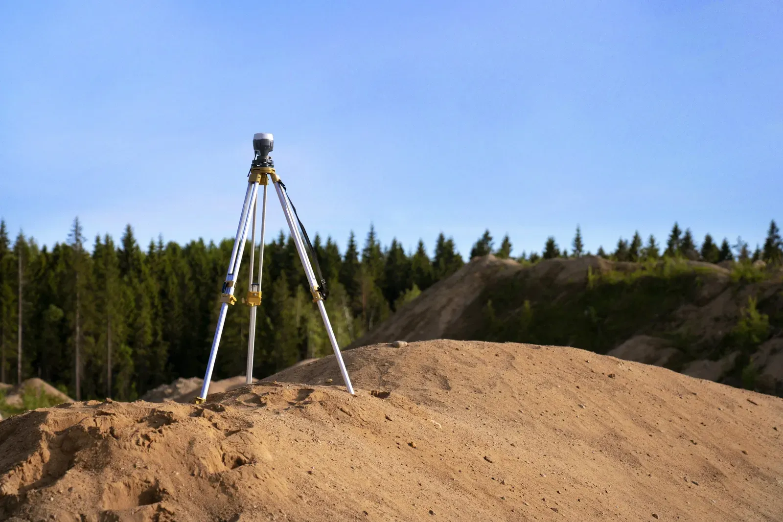 A GPS survey tripod set up on a gravel pile with a conifer forest in the background